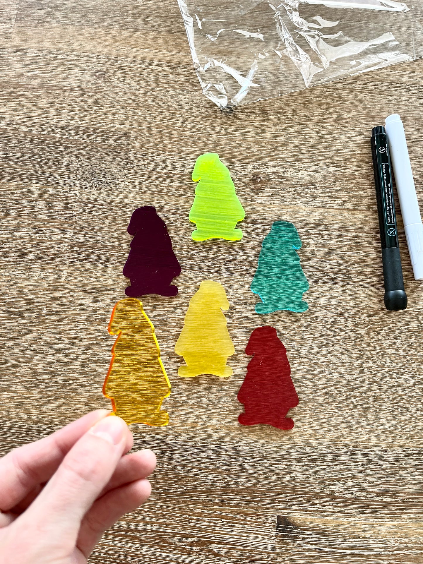 Hand holding a translucent yellow gnome-shaped piece with five additional colorful translucent gnomes and black and white dry erase markers on a wood surface, featuring Little Dry Erasables - Translucent Gnomes, handcrafted homeschool education resources.