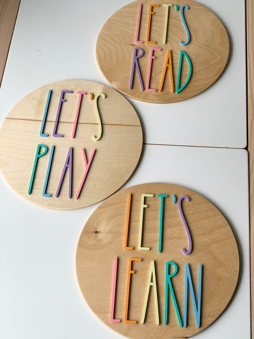 Set of three 10-inch round wooden signs with colorful pastel letters spelling Let’s Play, Let’s Read, and Let’s Learn