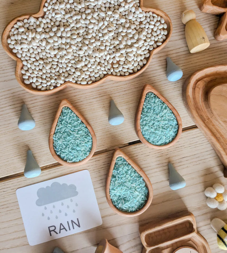 Cloud-shaped wooden sensory tray filled with white beans and raindrop-shaped trays with blue rice on wood table