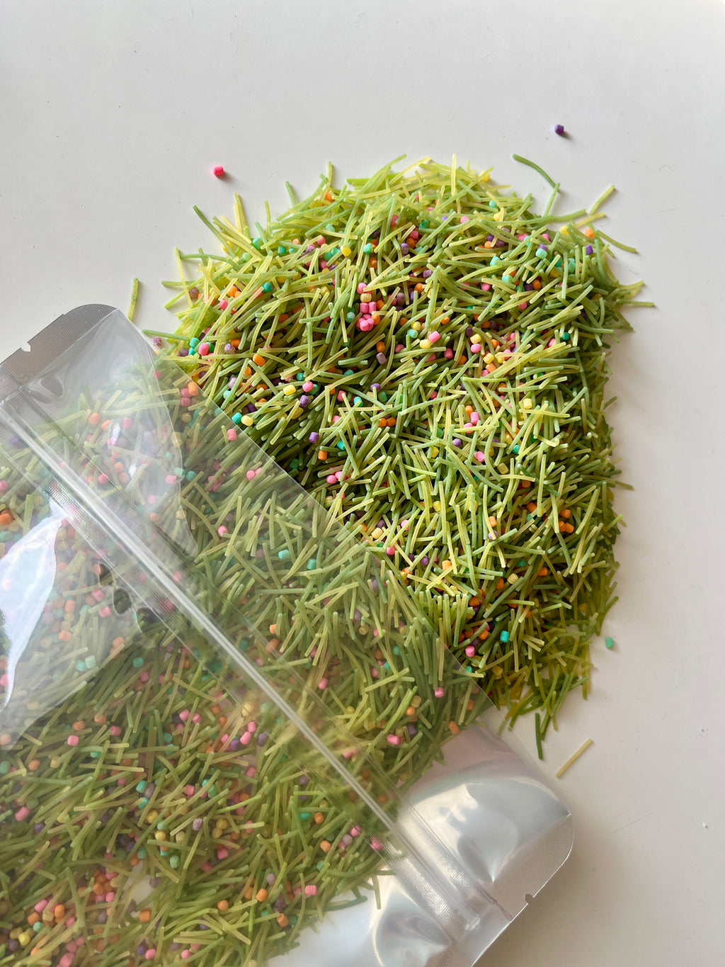 Clear resealable bag spilling thin green pasta strands mixed with tiny colorful round sprinkles on white surface