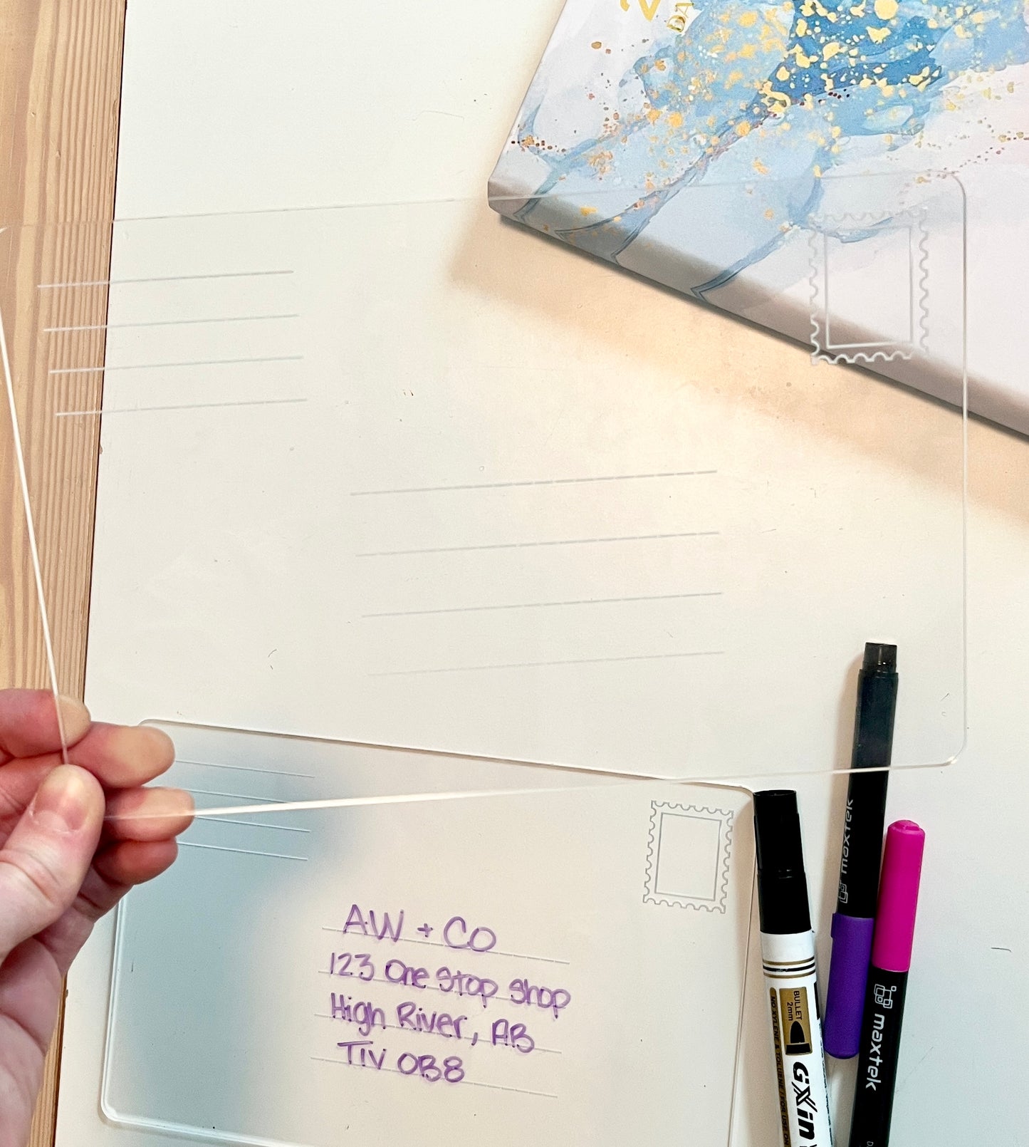 Hand holding a clear plastic sheet with writing, surrounded by stationery items on a white surface. Dry erase envelope for address practice