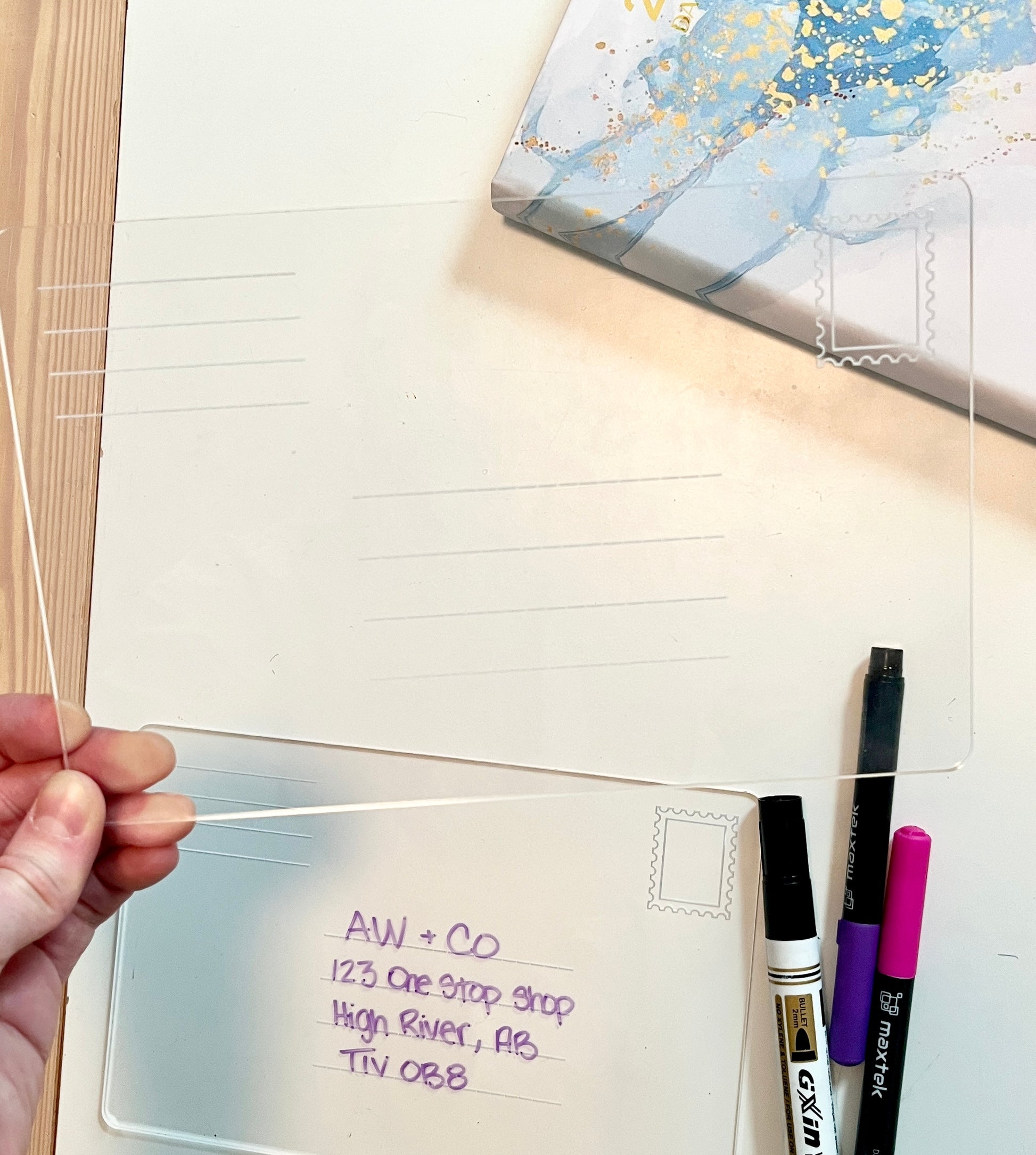 Hand holding a clear plastic sheet with writing, surrounded by stationery items on a white surface. Dry erase envelope for address practice