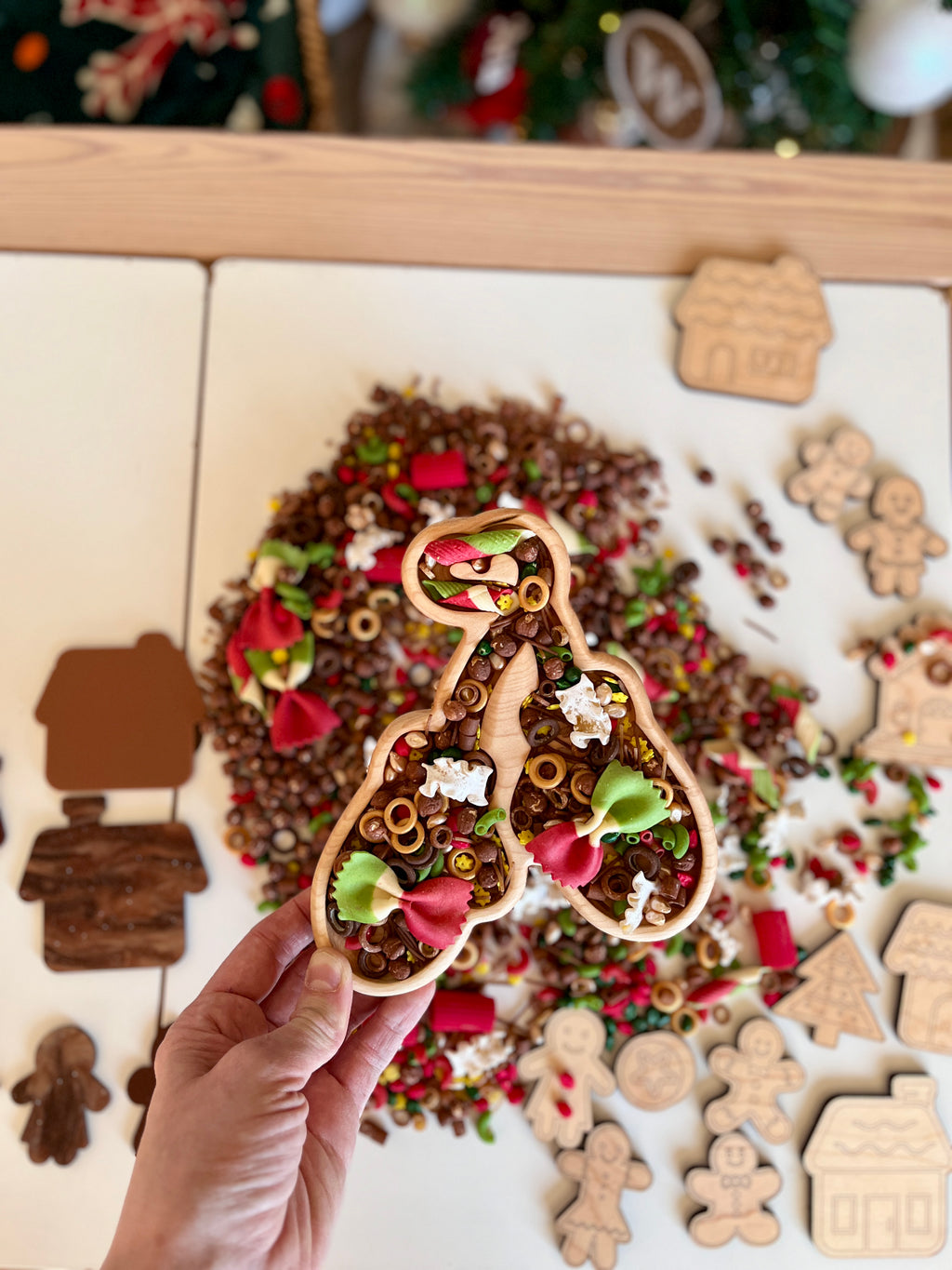 Hand holding wooden mitten-shaped tray filled with colorful pasta, beads, and small decorative holiday items on table