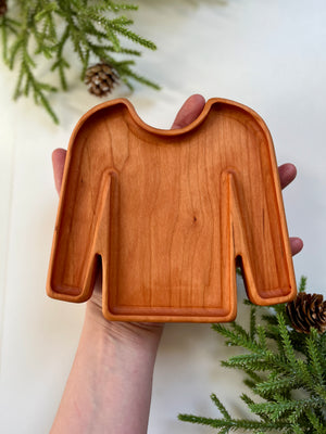 Hand holding a smooth wooden tray carved into the shape of a long-sleeve sweater with pine branches nearby