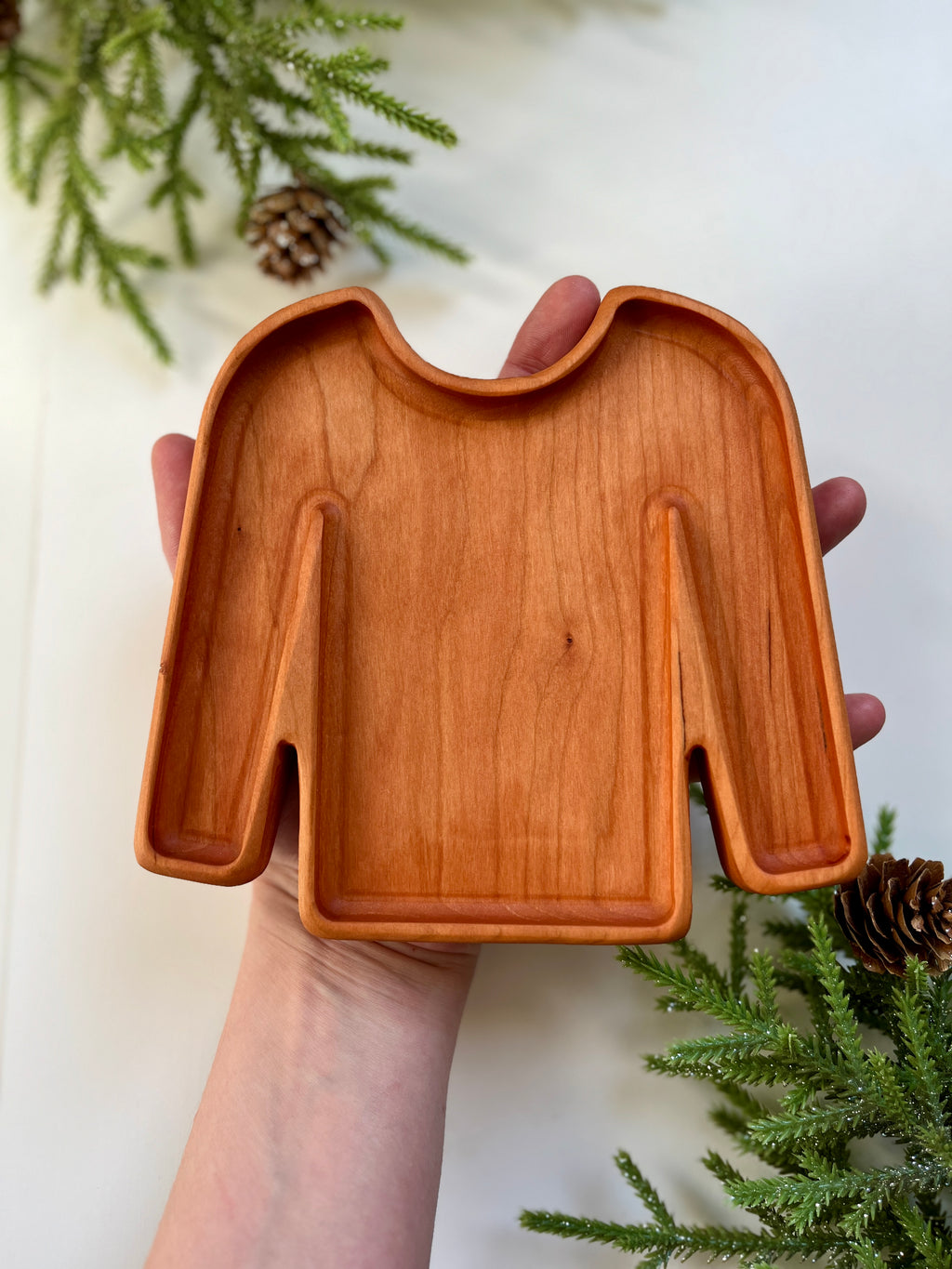 Hand holding a smooth wooden tray carved into the shape of a long-sleeve sweater with pine branches nearby