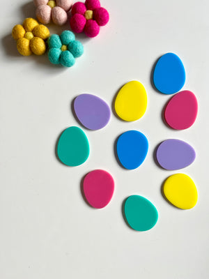Brightly colored plastic egg shapes arranged beside felt flowers on a white surface