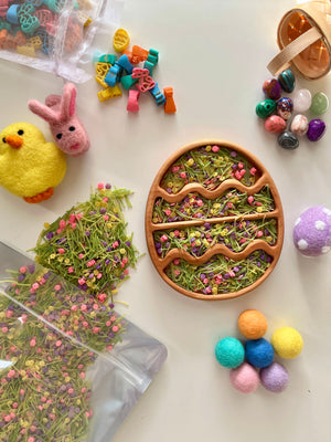 Wooden egg-shaped tray with dried green grass and pink flowers surrounded by colorful felt balls and decorative stones