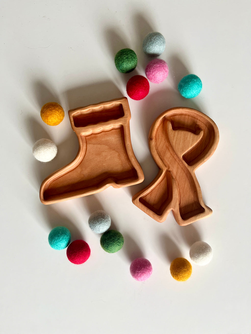 Two wooden trays shaped like a stocking and a ribbon surrounded by colorful felt balls on a white surface