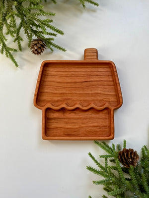 Wooden tray shaped like a house with chimney, surrounded by pine branches and pine cones on white surface