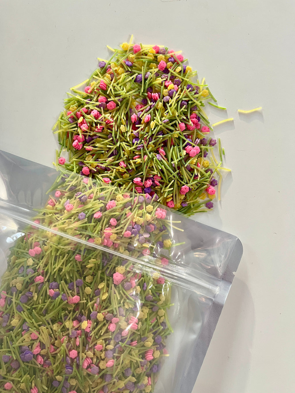 Clear resealable pouch spilling thin green pasta strands mixed with small pink and purple flower-shaped sprinkles on white background