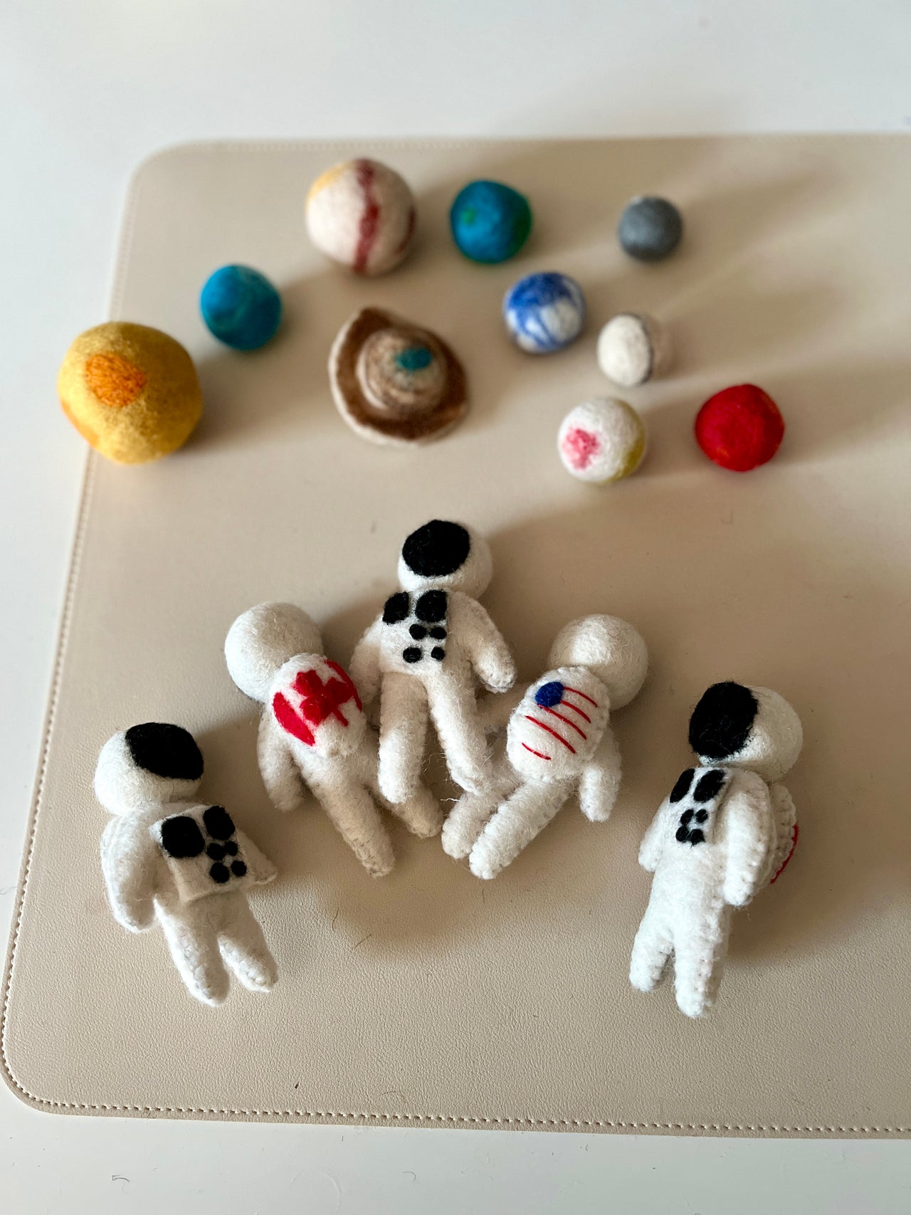 Five small handmade felt astronaut figures arranged on a beige surface with colorful felt planets in the background