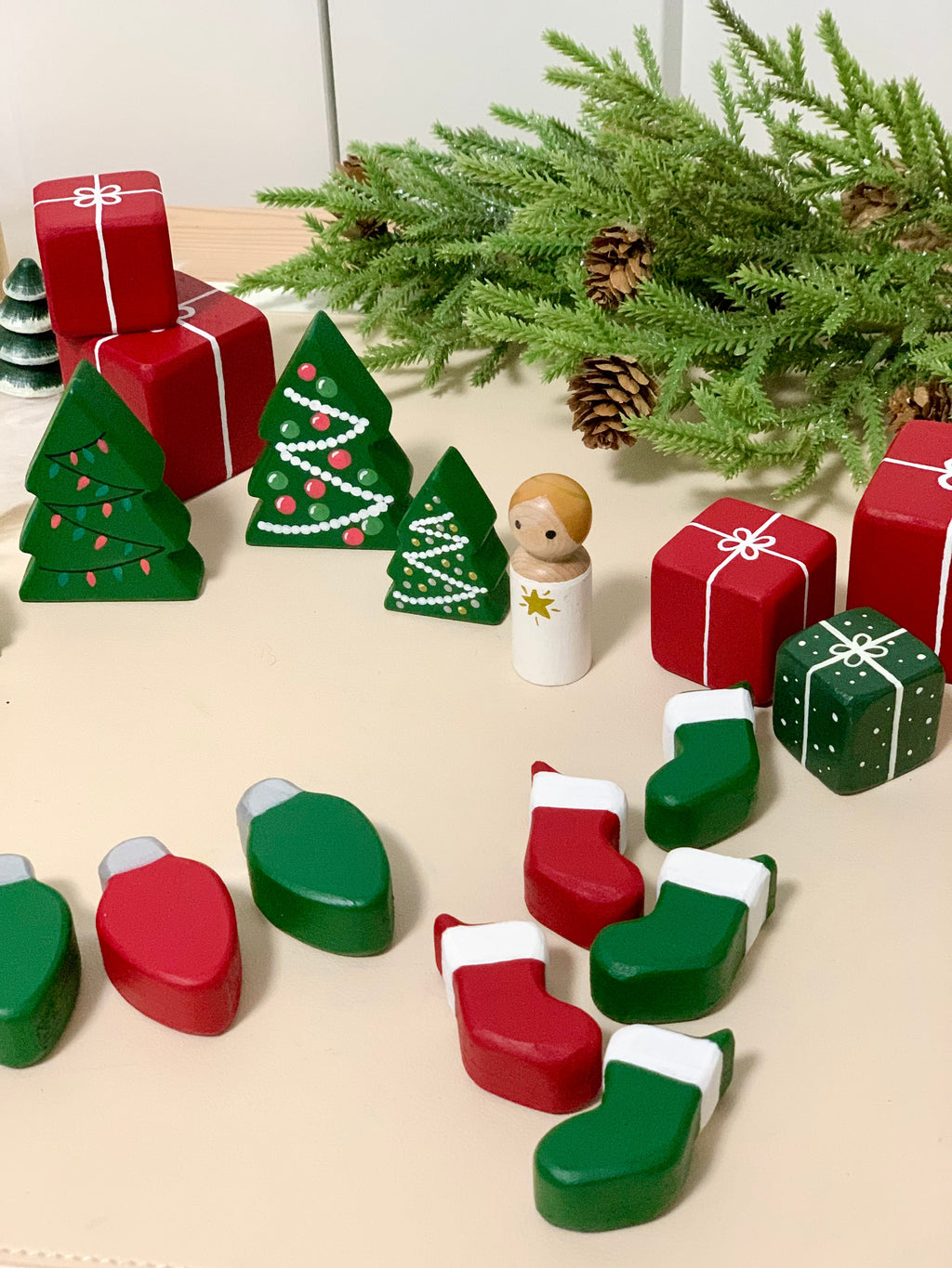 Set of painted wooden Christmas trees, presents, stockings, lights, and peg doll with pine branch background on beige surface