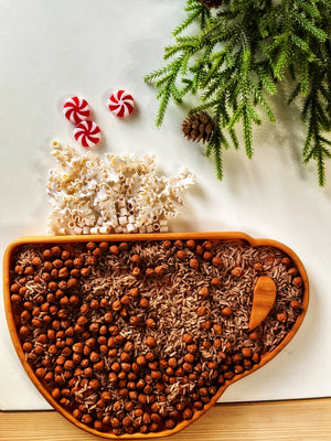 Wooden mitten-shaped tray filled with hazelnuts and rice, popcorn, peppermint candies, pine branches, and a pine cone on white surface