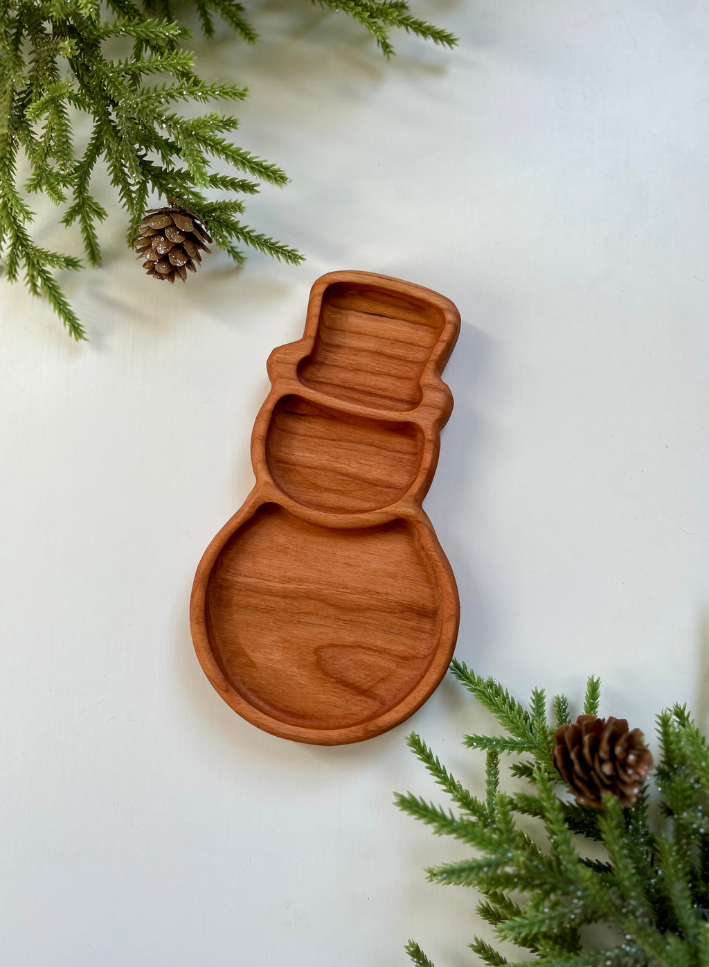 Carved wooden tray shaped like a snowman with sections, surrounded by pine branches and pine cones on white surface