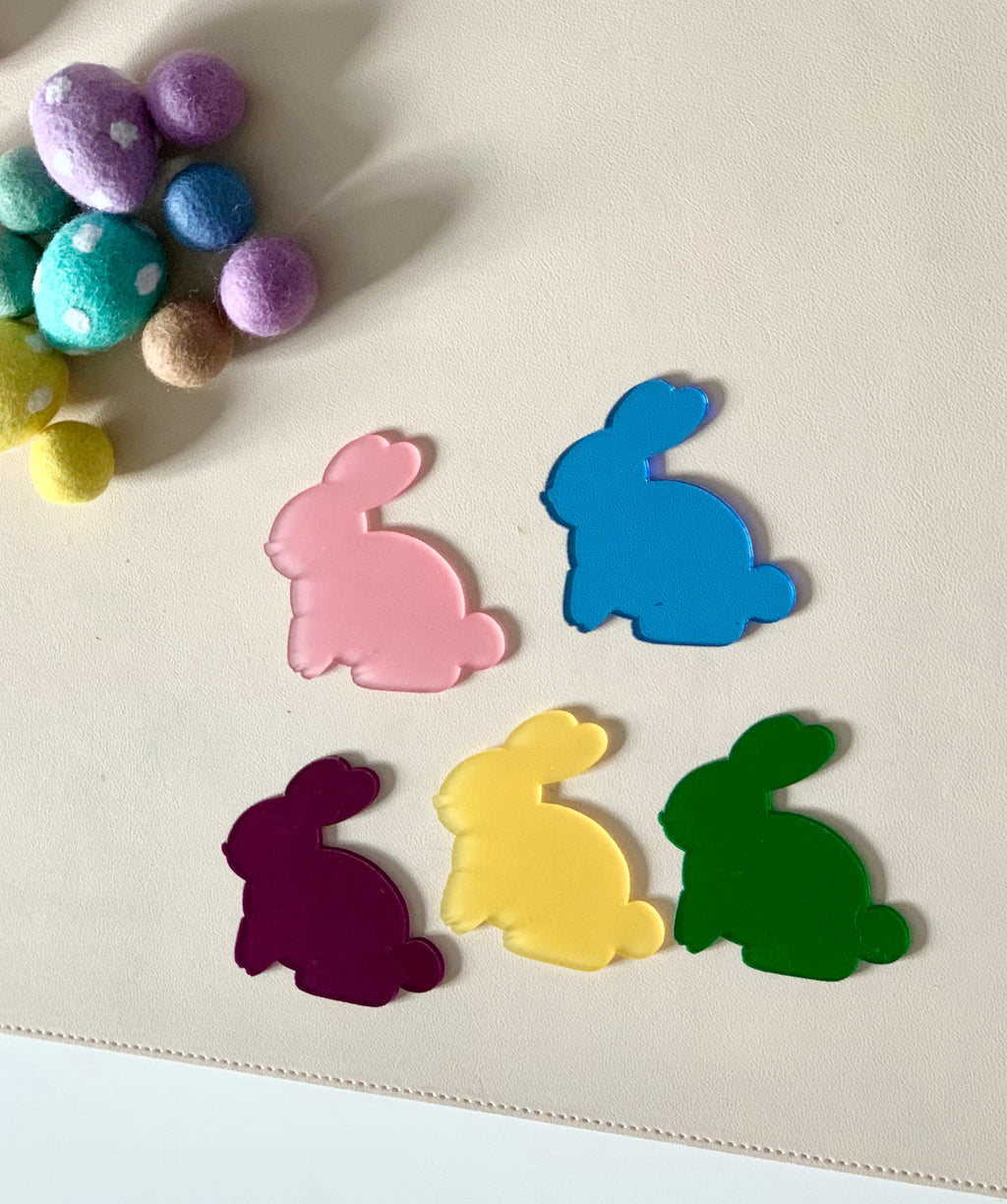 Five solid-colored bunny-shaped cutouts in pink, blue, purple, yellow, and green on a light surface with felt balls nearby