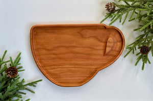 Wooden serving tray shaped like a coffee cup with a carved handle on white surface with pine branches and cones
