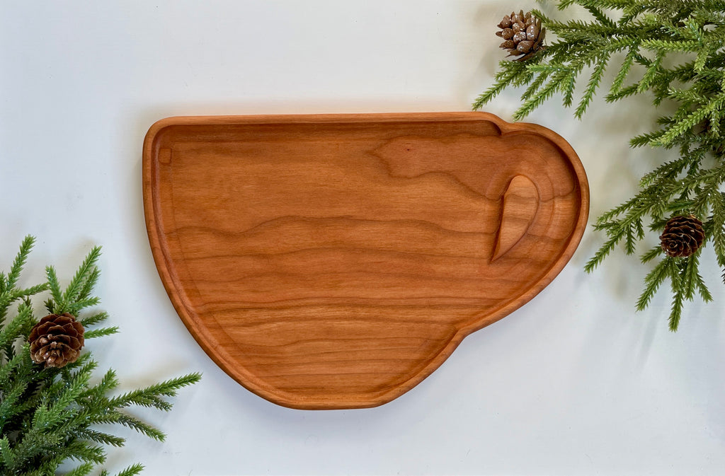 Wooden serving tray shaped like a coffee cup with a carved handle on white surface with pine branches and cones