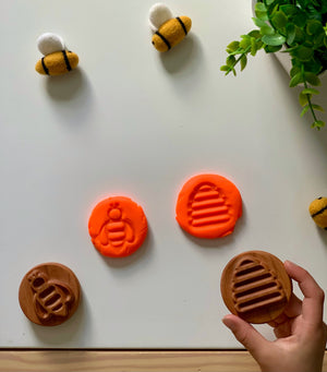 Hand holding round wooden stamp with beehive design, matching orange clay impressions, felted bee toys, and green plant on white surface