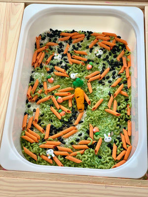 White container filled with orange pasta, green spiral pasta, black beans, rice, and small bunny and carrot decorations