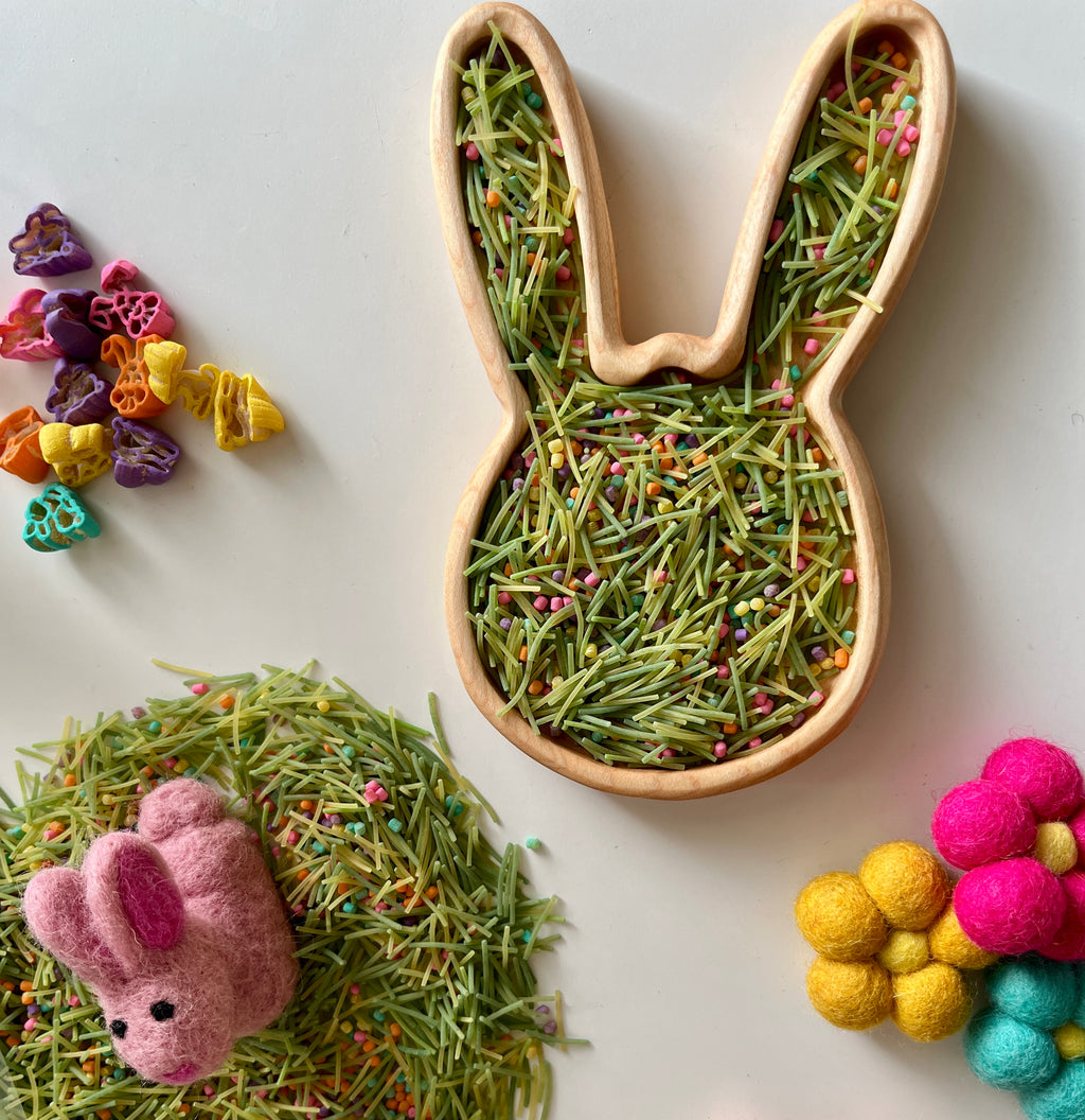 Wooden bunny-shaped tray filled with green pasta and colorful sprinkles beside felt rabbit and pom-pom flower decorations