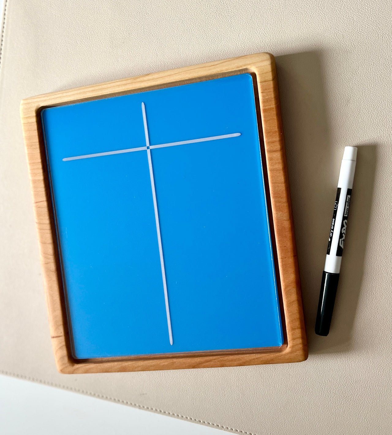 Rectangular wooden frame with blue dry erase board showing a white cross and a black dry erase marker on beige surface