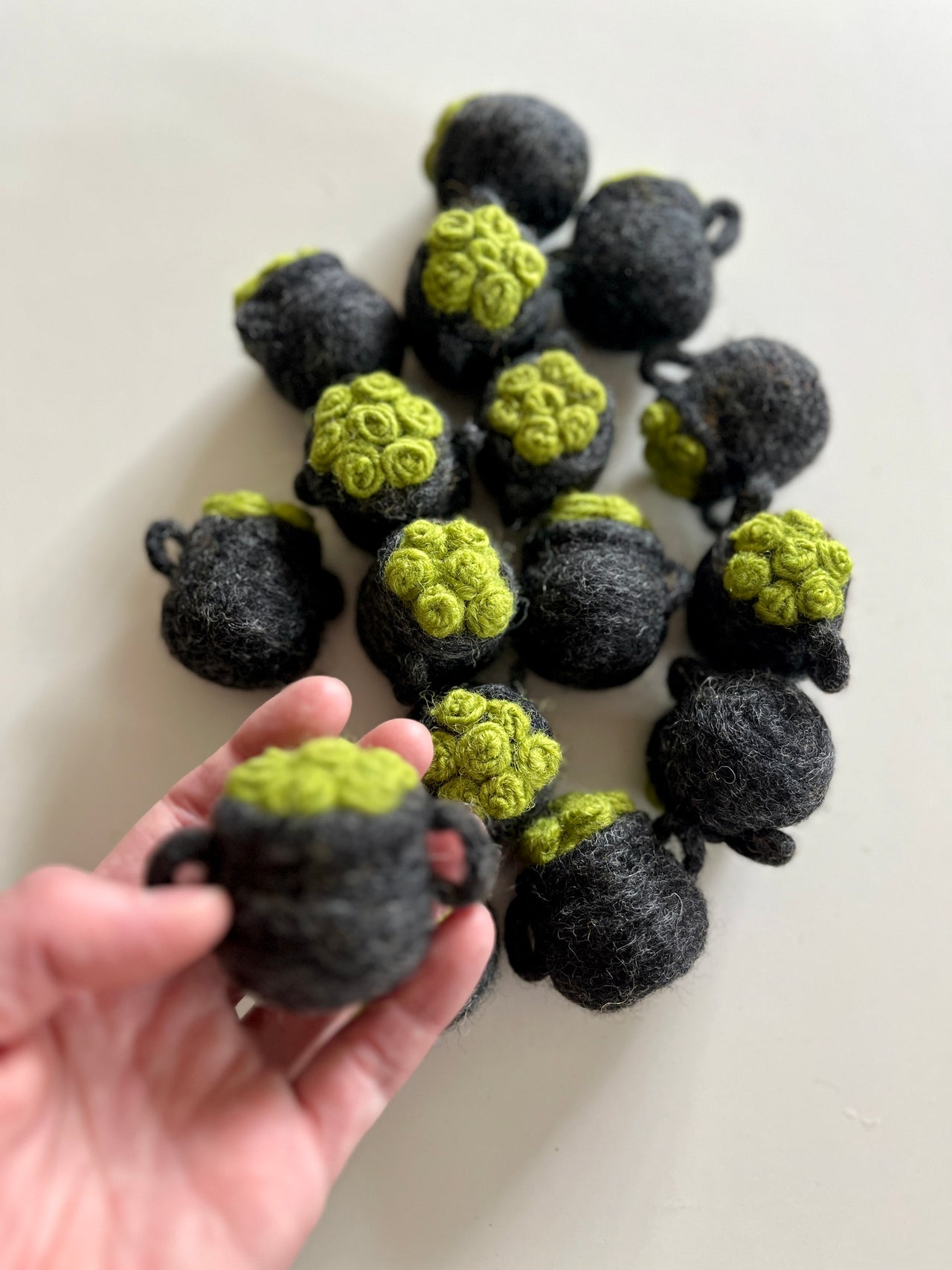 Hand holding one small dark gray felt pot with bright green textured top, with several similar pots scattered on white surface.