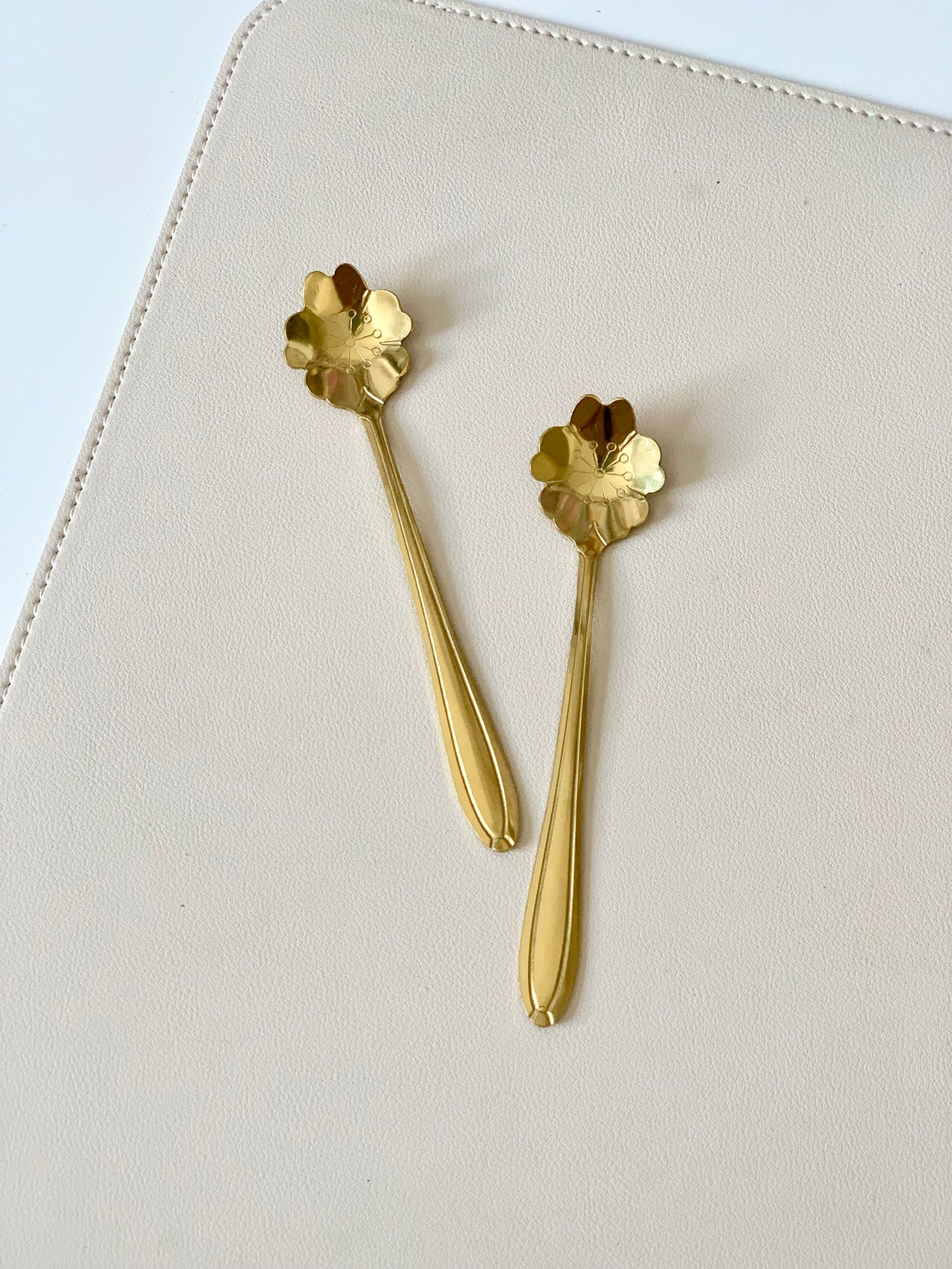 Two gold metal spoons with scalloped flower-shaped bowls on a light beige textured surface