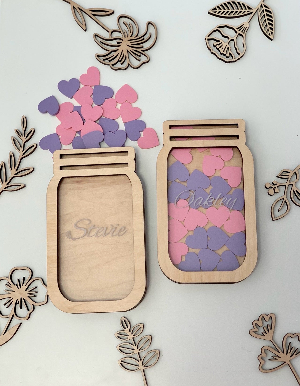 Two personalized wooden jar-shaped frames with pink and purple heart tokens surrounded by wooden floral cutouts on white surface