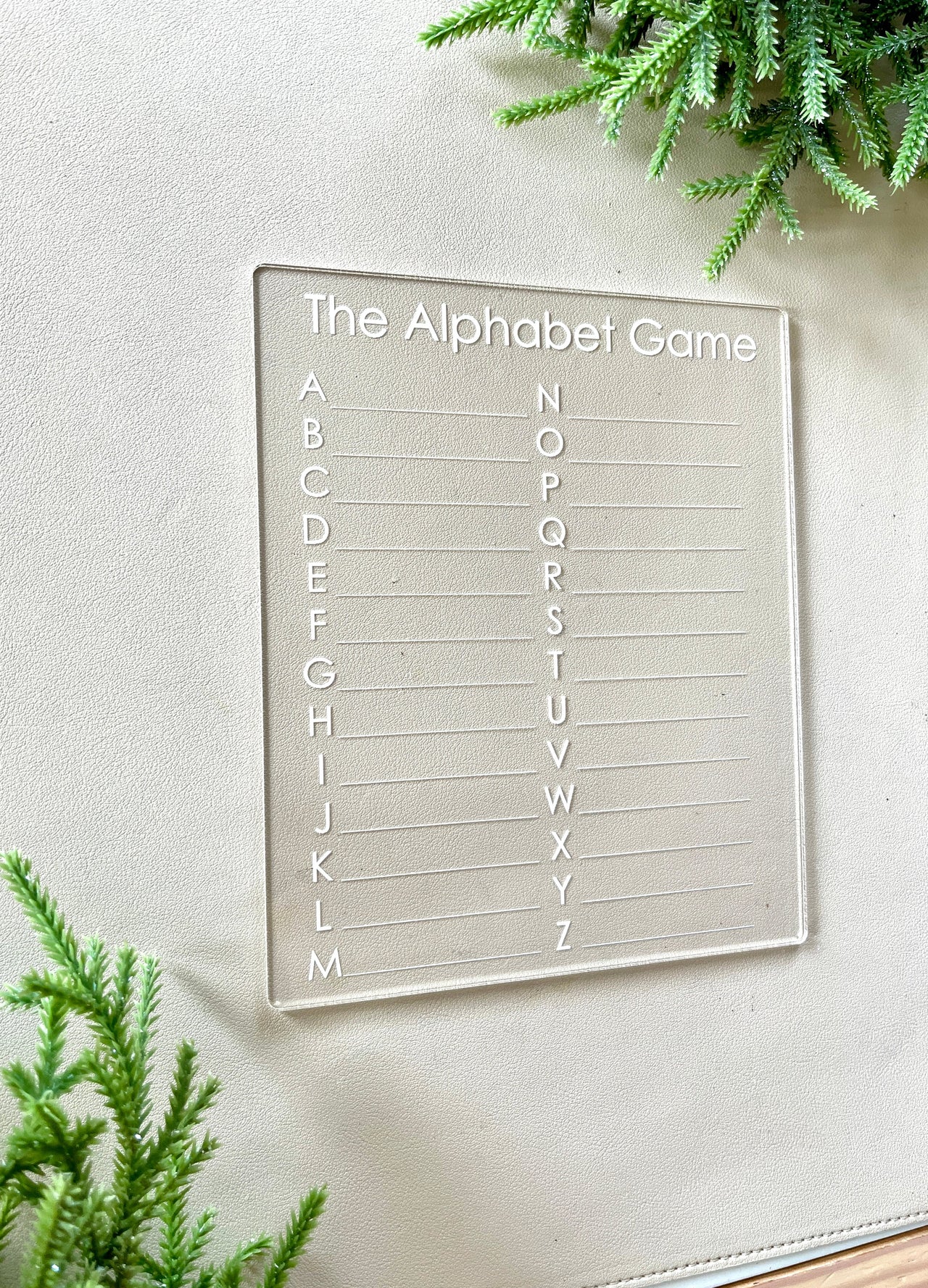 Clear acrylic game board engraved with the alphabet from A to Z and labeled "The Alphabet Game" on a light surface with greenery