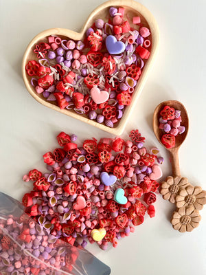 Heart-shaped wooden bowl filled with red, pink, and purple pasta shapes and pastel heart-shaped beads on white surface