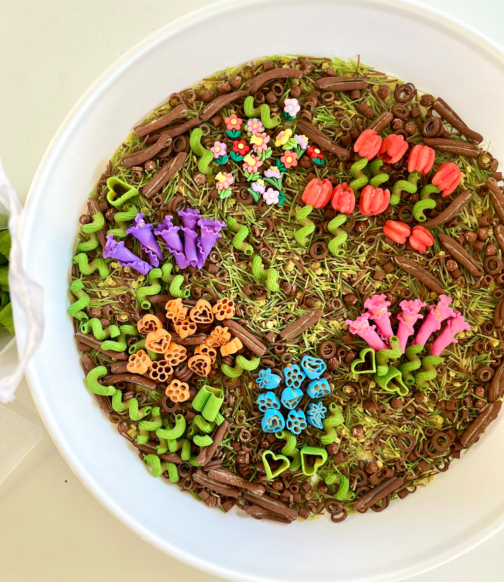 Round white bowl filled with colorful floral-shaped pasta and green spirals on dried grass and brown pasta mix
