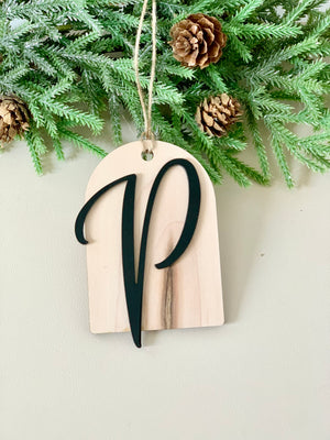 Light natural wood arch ornament with a black cursive letter V hanging from twine against pine branches and cones