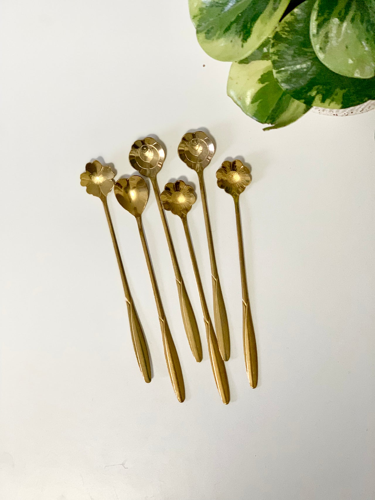 Six gold-toned spoons with long handles and flower-shaped bowls arranged on a white surface near green leaves
