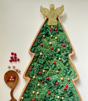 Tree-shaped wooden tray filled with green shredded paper, red and gold beads, and topped with a glittery gold angel ornament