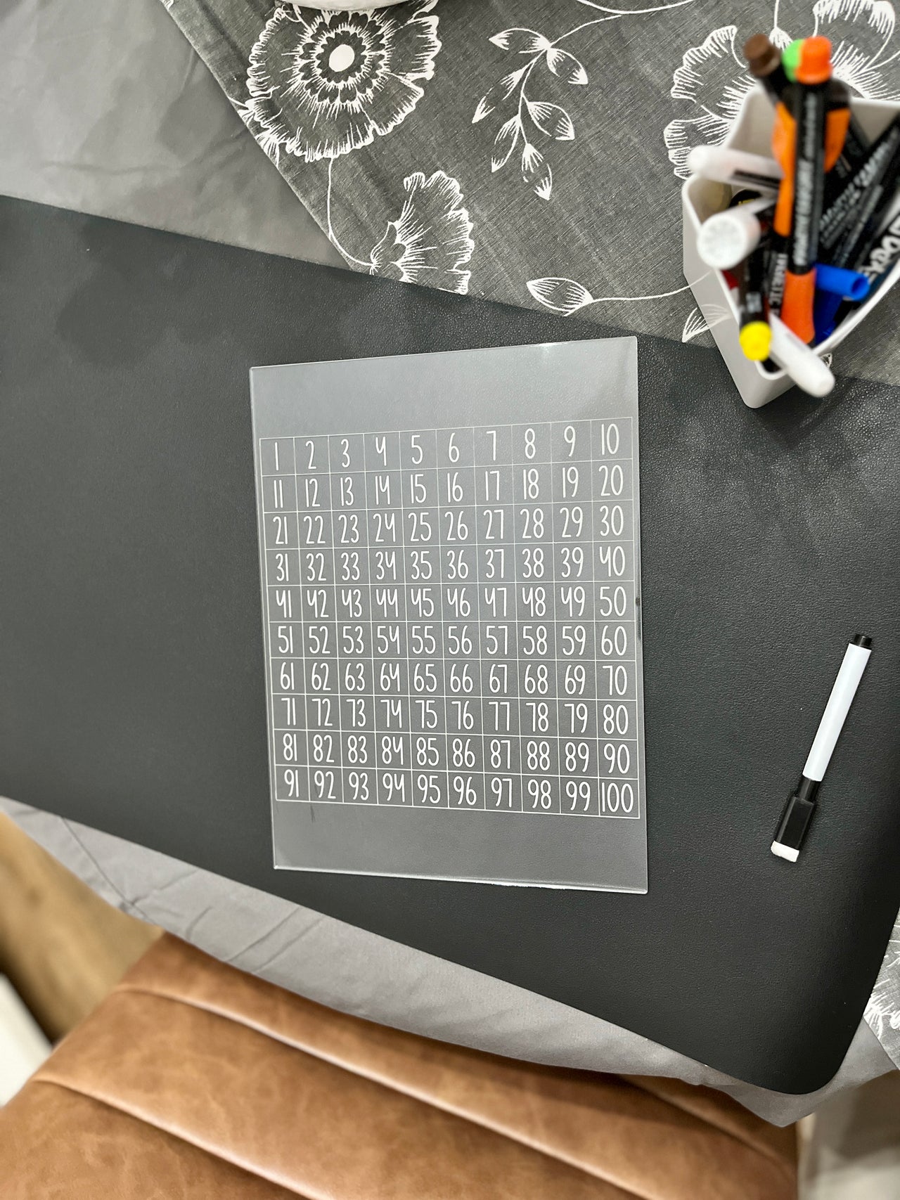 Clear acrylic sheet with numbered grid 1 to 100 beside a black dry erase marker and container of colorful markers on table