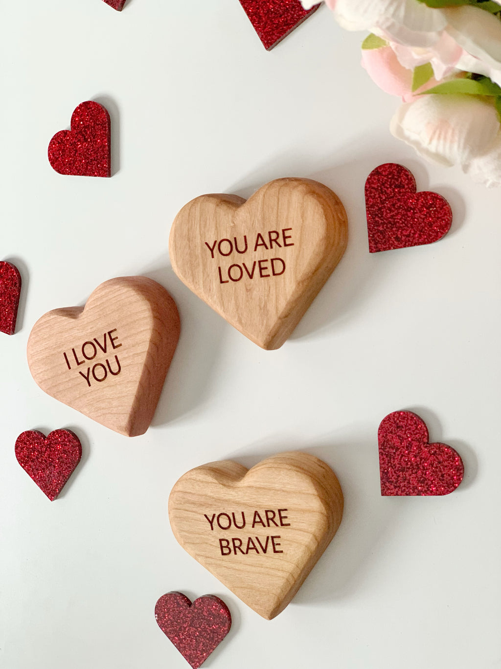 Three wooden heart-shaped blocks engraved with positive messages surrounded by small red glitter hearts on white surface