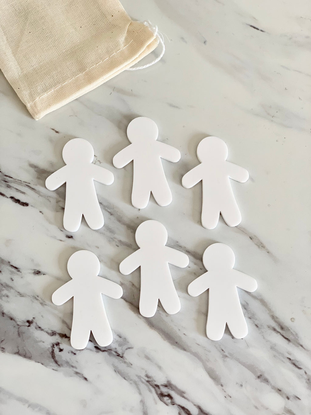 Six small white dry erase people shapes arranged on marble surface with beige drawstring bag nearby