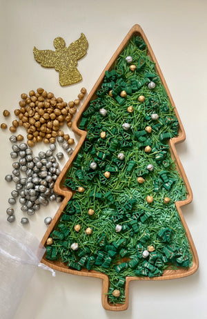 Wooden tray shaped like a Christmas tree filled with green shredded paper and scattered silver and gold beads next to metallic beads and glittery angel cutout