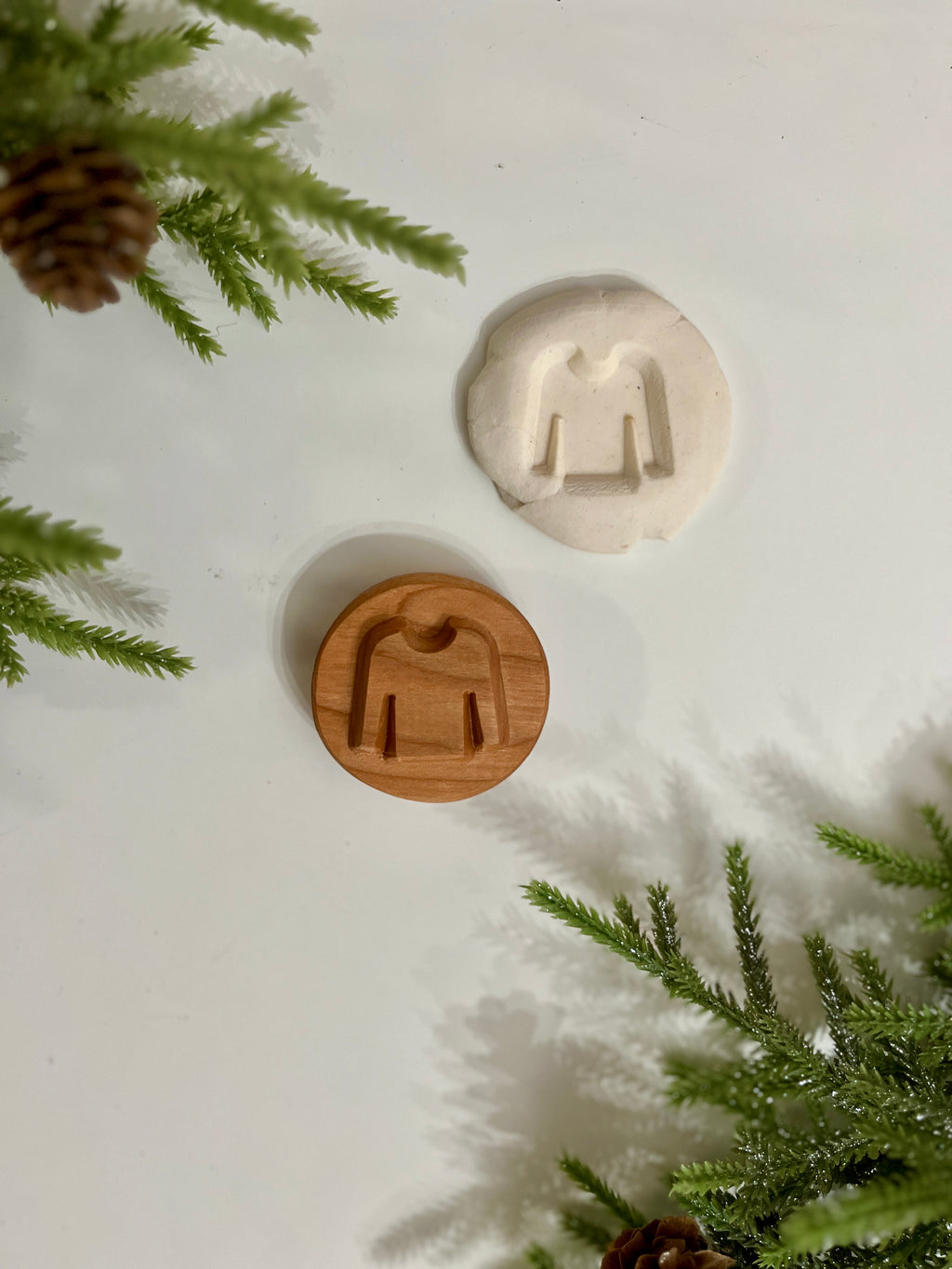 Round wooden cookie stamp with sweater design and matching white dough impression on a white surface with green pine branches