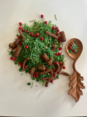 Wooden oak leaf-shaped spoon with dried green herbs, small red berries, and brown seed pods on white background