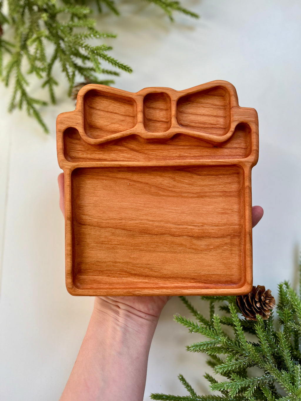 Hand holding a wooden tray carved in the shape of a gift box with a bow, surrounded by pine branches and pine cones