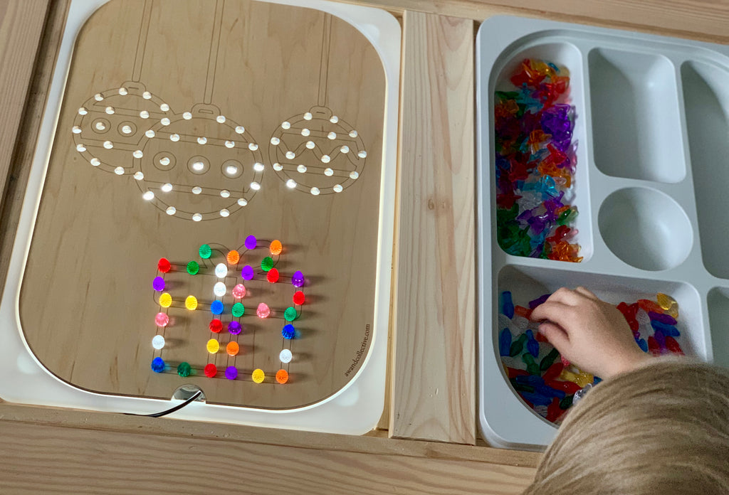 Wooden Flisat insert with holes for colorful pegs forming Christmas ball and gift shapes beside trays of translucent pegs and child's hand