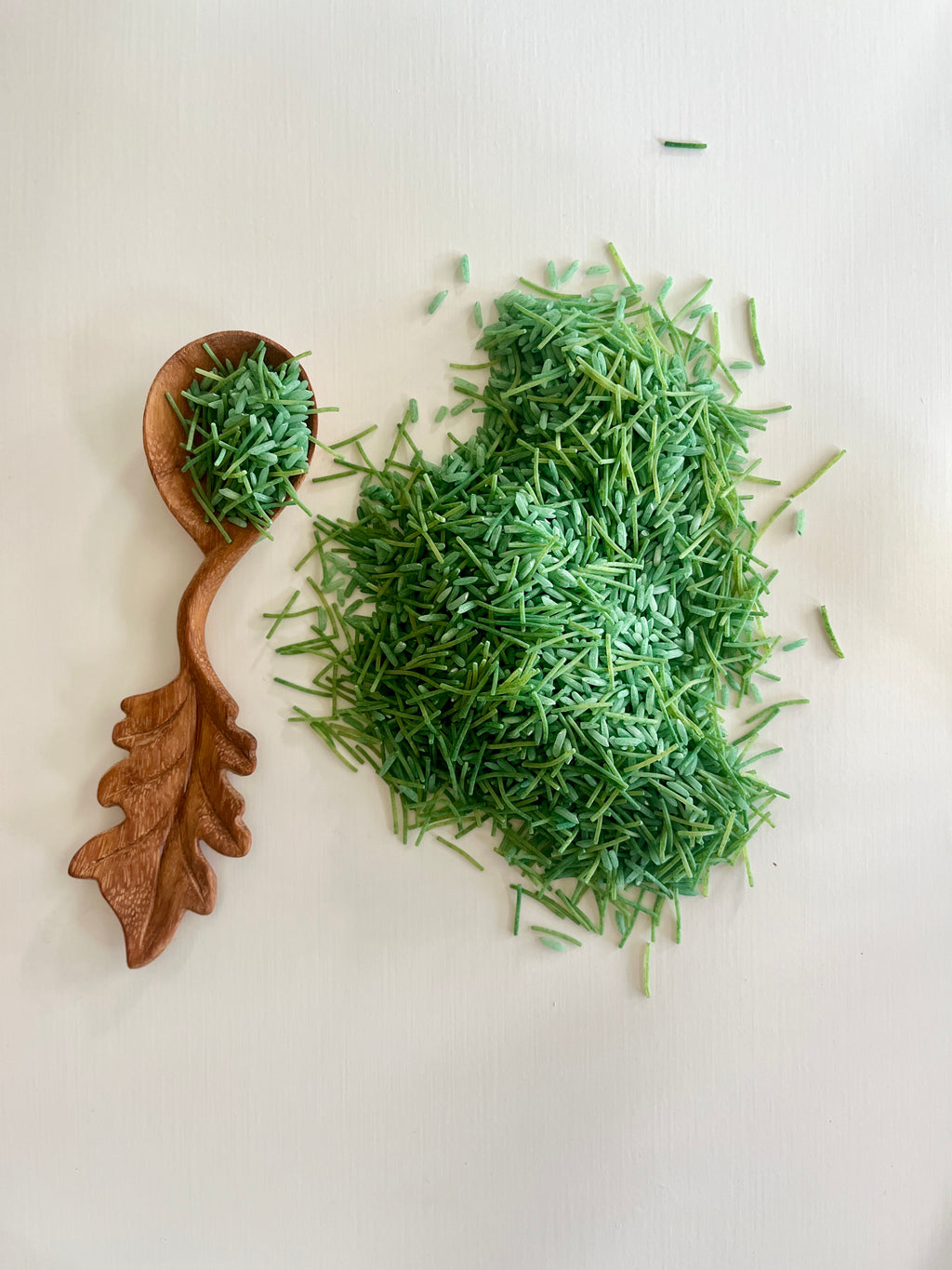 Carved wooden spoon shaped like an oak leaf holding green dried herbs next to a scattered pile of the same herbs on white background