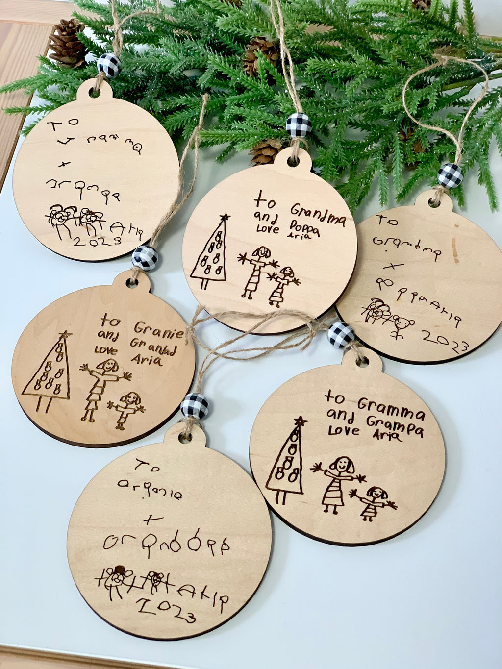 Six round wooden holiday ornaments with childlike drawings and handwritten messages hanging on twine near greenery and pine cones