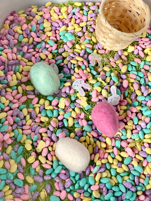 Small wooden basket resting on colorful pastel sensory beans with felt Easter eggs and bunny and chick decorations