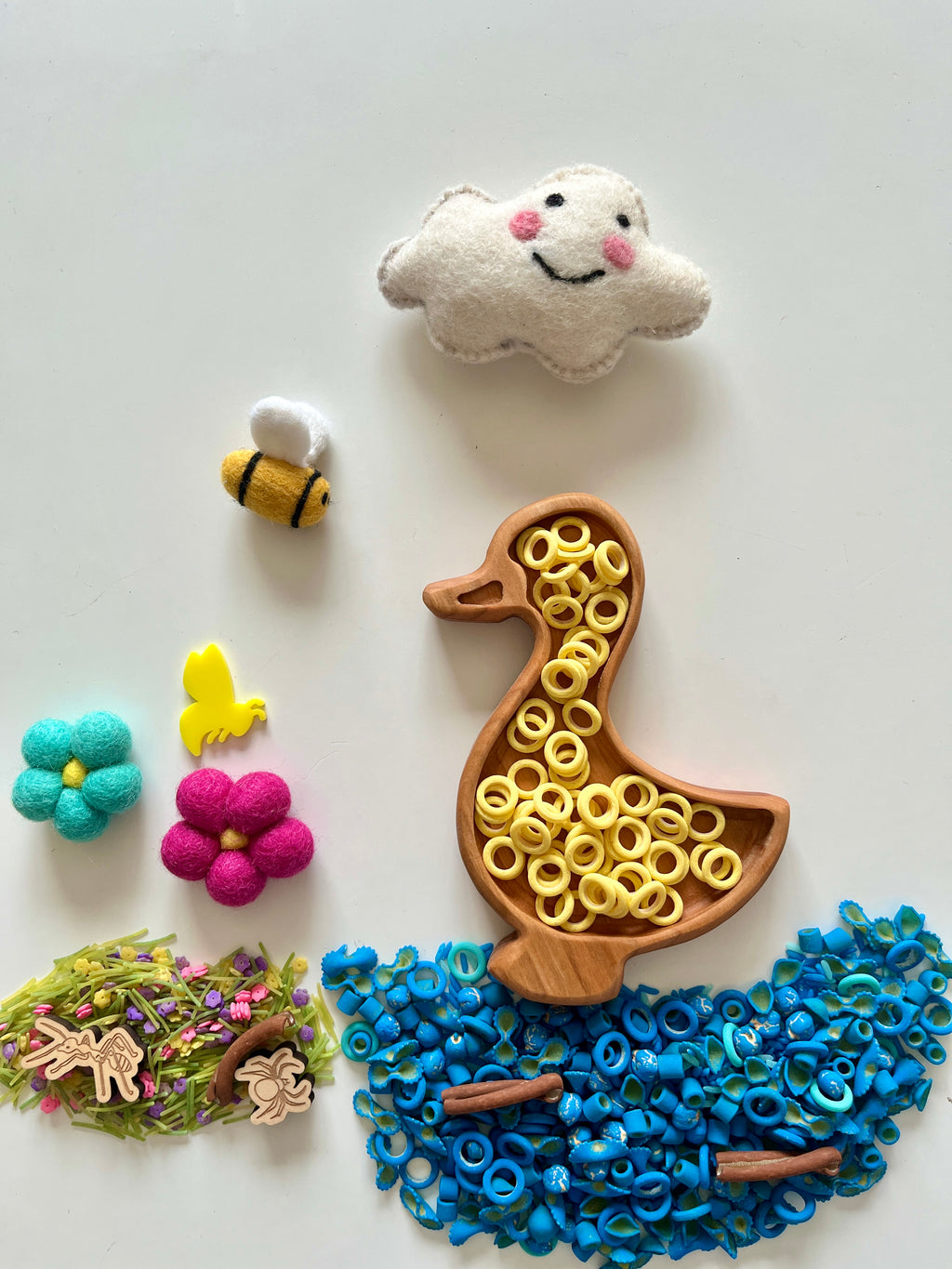 Wooden duck-shaped tray filled with yellow ring pasta on white surface surrounded by blue pasta rings, felt flowers, bees, and a smiling cloud
