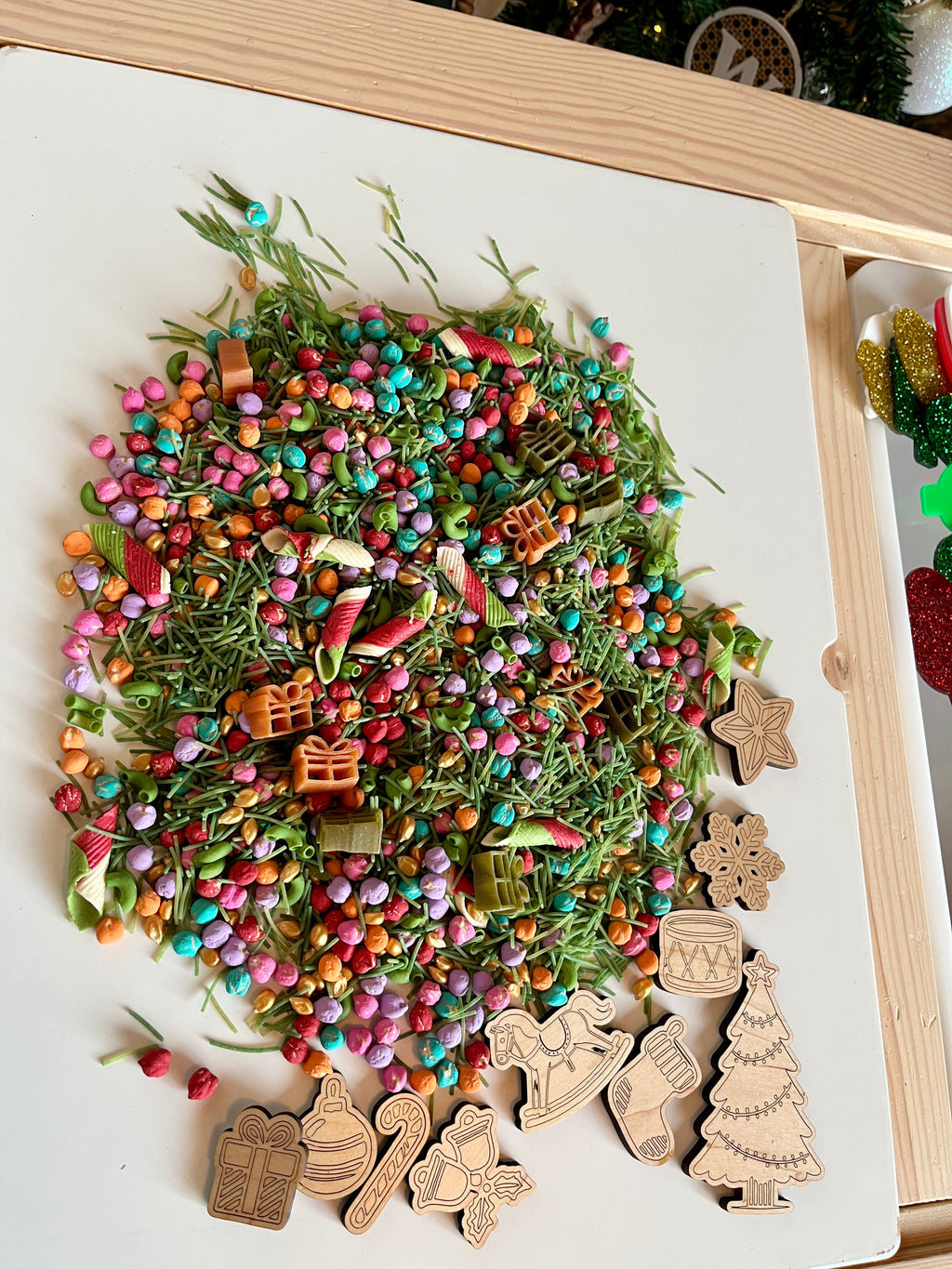 Colorful mix of dried greenery, small beads, pasta shapes, and wooden Christmas-themed cutouts on white tray