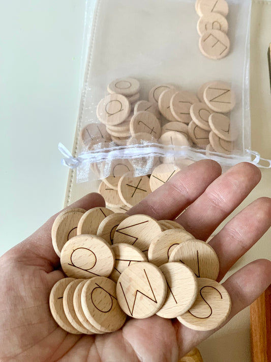 Hand holding several 1-inch natural wooden discs with laser engraved alphabet letters next to mesh pouch with more discs