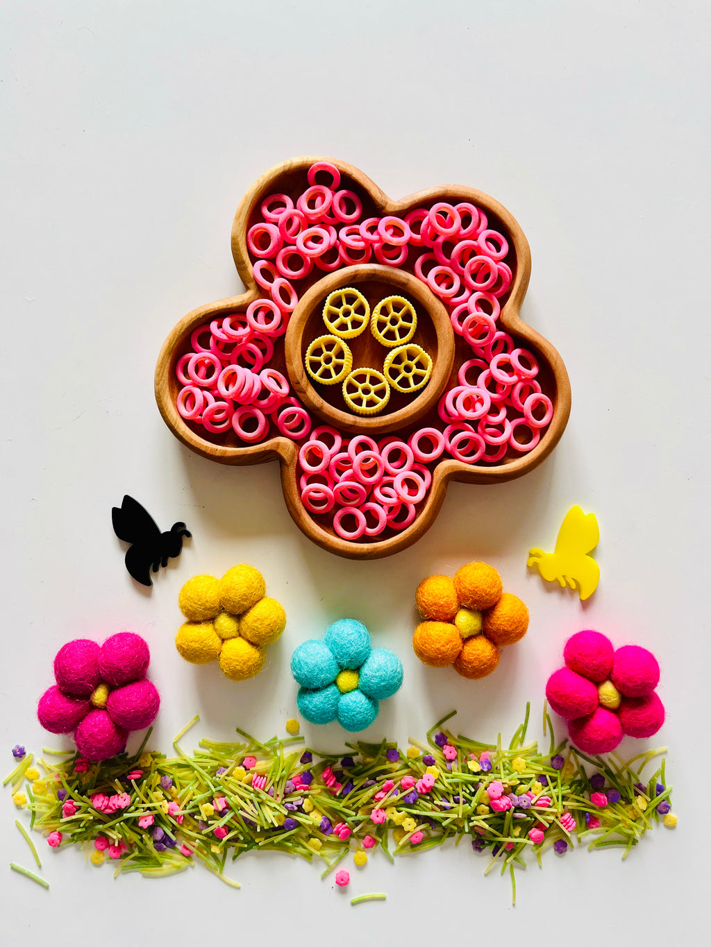 Flower-shaped wooden tray filled with pink rings and yellow wheel-shaped pasta with colorful felt flowers and bee cutouts below