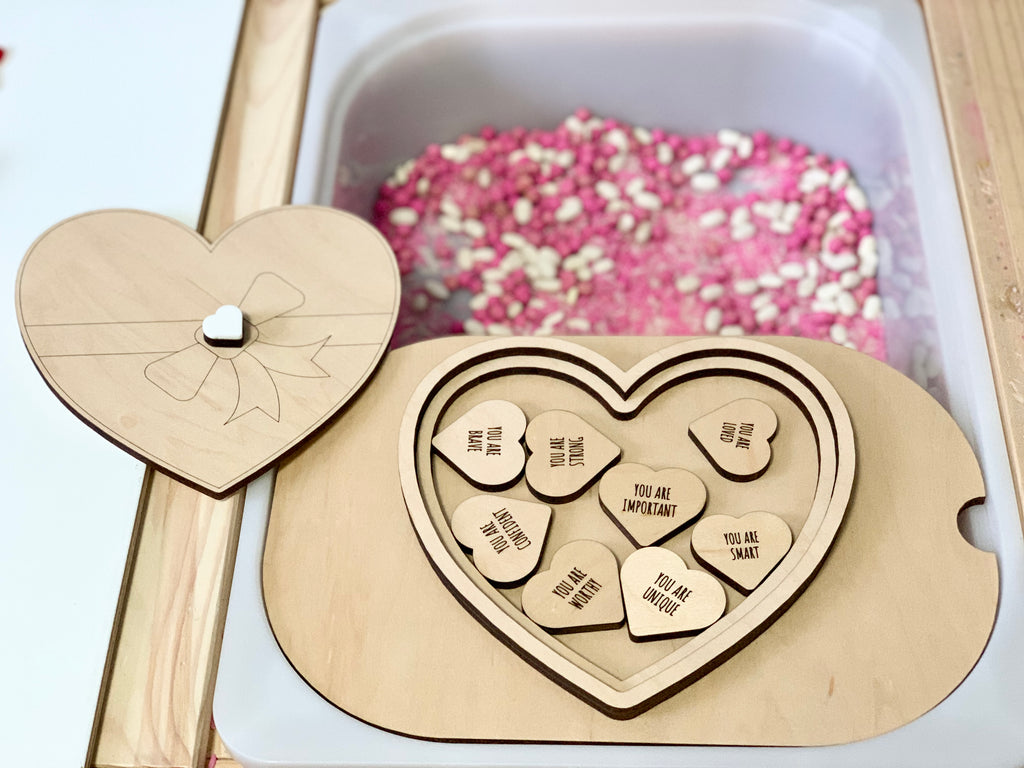 Heart-shaped wooden puzzle box with engraved positive affirmations next to a container of pink and white sensory beads